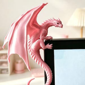 Pode incluir: Uma estatueta de drag&atilde;o rosa claro pousada no canto superior da moldura de um monitor preto. O drag&atilde;o apresenta escamas detalhadas, grandes asas abertas e uma cauda longa e curva que pende pela lateral da tela como um detalhe decorativo.