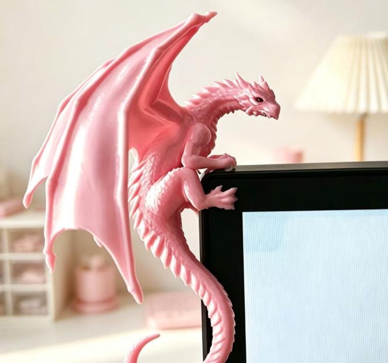 Pode incluir: Uma estatueta de drag&atilde;o rosa claro pousada no canto superior da moldura de um monitor preto. O drag&atilde;o apresenta escamas detalhadas, grandes asas abertas e uma cauda longa e curva que pende pela lateral da tela como um detalhe decorativo.