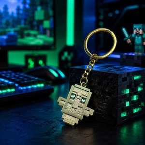 Pode incluir: Um porta-chaves de metal dourado com uma cabeça de personagem pixelizada e quadrada, com uma superfície texturizada e dois pequenos olhos quadrados verdes incrustados. O pendente está preso a um anel de metal por uma corrente curta.