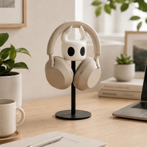 May include: A beige over-ear headphone set resting on a white, horned character-shaped stand. The stand features a smooth, matte white head with two prominent horns and black oval eye sockets, supported by a thin black vertical rod and a round base.