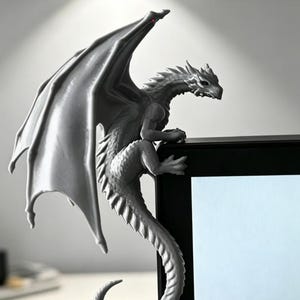 Pode incluir: Uma estatueta de drag&atilde;o cinzento pousada no canto superior de um monitor preto. O drag&atilde;o tem asas grandes e estendidas, um corpo texturizado com escamas e uma cauda longa que se curva para baixo ao longo da lateral do ecr&atilde;.
