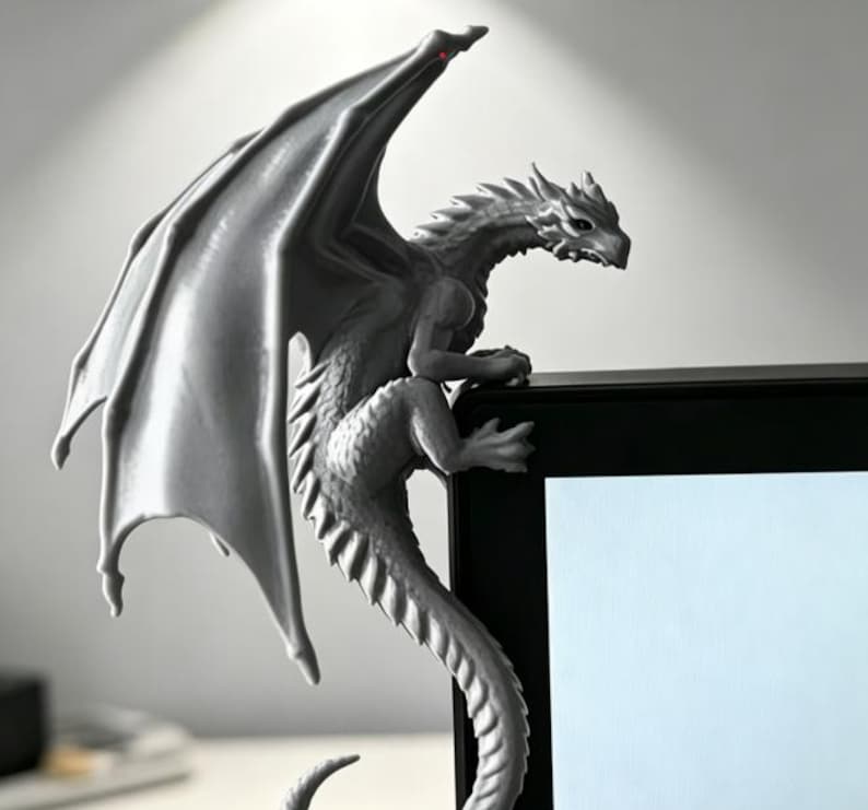 Pode incluir: Uma estatueta de drag&atilde;o cinzento pousada no canto superior de um monitor preto. O drag&atilde;o tem asas grandes e estendidas, um corpo texturizado com escamas e uma cauda longa que se curva para baixo ao longo da lateral do ecr&atilde;.
