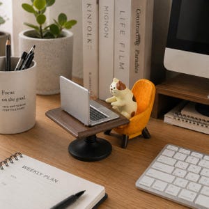 May include: A miniature figurine of a calico cat sitting in a small orange armchair at a dark brown wooden desk with a silver laptop. The cat has white, brown, and black markings, and its paws are positioned as if typing on the laptop keyboard.