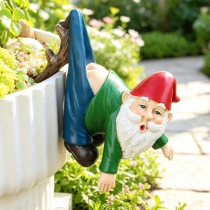 Puede incluir: Una figura de gnomo de jardín colgada del borde de una maceta blanca. El gnomo lleva un gorro rojo, una camisa verde y pantalones azules, con los pantalones bajados para mostrar sus glúteos. Tiene una barba blanca y expresión de sorpresa.
