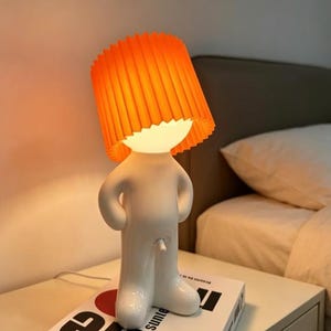 Pode incluir: Um candeeiro de mesa branco em forma de figura humana estilizada com as mãos na cintura. O candeeiro apresenta um abajur cilíndrico plissado de cor laranja brilhante que assenta sobre a cabeça, emitindo uma luz quente e suave.