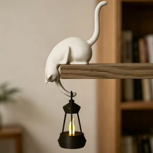 Pode incluir: Uma estatueta de gato branco pousada na borda de uma prateleira de madeira com a cauda pendurada. A cauda do gato sustenta uma pequena lanterna preta contendo uma luz amarela brilhante em forma de chama, criando uma peça decorativa suspensa.
