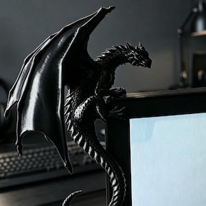 Pode incluir: Uma estatueta de drag&atilde;o preto fosco projetada para se empoleirar na borda superior de um monitor de computador. A criatura apresenta escamas detalhadas, grandes asas texturizadas estendidas para cima e uma cauda longa que curva na lateral.
