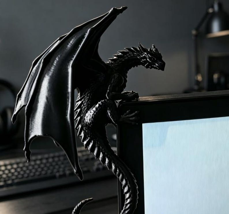 Pode incluir: Uma estatueta de drag&atilde;o preto fosco projetada para se empoleirar na borda superior de um monitor de computador. A criatura apresenta escamas detalhadas, grandes asas texturizadas estendidas para cima e uma cauda longa que curva na lateral.