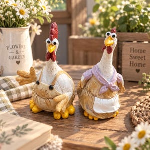 Könnte beinhalten: Zwei Hühnerfiguren mit Wackelaugen. Das linke Huhn trägt eine hellbraune Latzhose, einen roten Kamm und einen gelben Schnabel. Das rechte Huhn trägt ein lavendelfarbenes Tuch und eine weiße Schürze. Beide haben texturierte weiß-braune Federn und gelbe Füße.