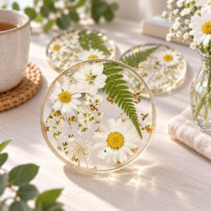Könnte beinhalten: Ein Set aus drei runden Untersetzern aus Kunstharz mit eingebetteten weißen Gänseblümchen, grünen Farnblättern und kleinen weißen Füllblumen. Jeder Untersetzer ist mit kleinen Goldfolienpartikeln im klaren, glatten und transparenten Harzmaterial verziert.