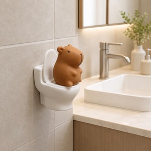 Peut inclure: Une figurine de capybara marron assise dans un petit support blanc en forme de toilettes. Le capybara a un corps arrondi, de petites oreilles et une expression faciale simple. Le support blanc possède un couvercle relevé et est fixé au mur.