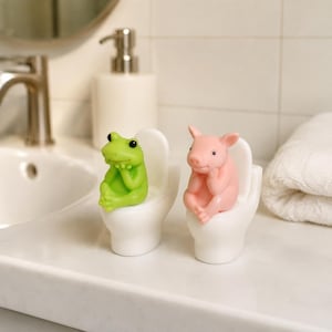 Può includere: Due piccole statuette, una rana verde e un maialino rosa, sedute ciascuna su un gabinetto in miniatura bianco. Le figure hanno le mani appoggiate sotto il mento in una posa pensierosa. Gli oggetti sono lisci, opachi e di materiale solido.