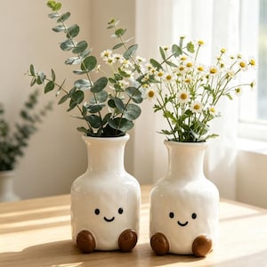 May include: Two white ceramic vases with a textured surface, each featuring a simple black painted smiley face and two brown rounded feet at the base. One vase holds green eucalyptus stems, while the other contains white daisies with yellow centres.