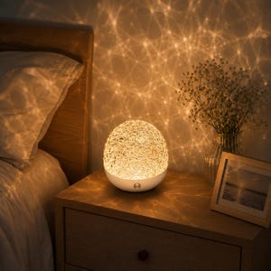 Peut inclure: Une lampe de table sphérique avec un dôme supérieur translucide et texturé qui émet une lumière chaude à motifs. La base est en matériau blanc uni et comporte un petit bouton d'alimentation métallique circulaire au centre de l'hémisphère inférieur.