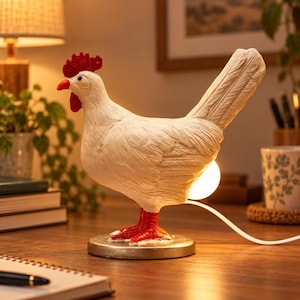 Pode incluir: Um candeeiro de mesa em forma de galinha branca com crista e barbelas vermelho-vivo, assente numa base circular prateada. O candeeiro apresenta um corpo texturado e uma lâmpada na parte traseira que emite uma luz quente através de um cabo branco.