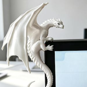 May include: A white dragon figurine with detailed scales, large wings, and a long tail, designed to perch on the top corner of a computer monitor. The creature has a sharp, angular head and claws gripping the edge of the black screen frame.