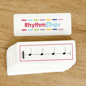 May include: A stack of white rectangular cards featuring musical notation. The top card displays the text RhythmStrips in red and blue with colorful dashes. The other cards show a 4/4 time signature with four quarter notes in a red border.