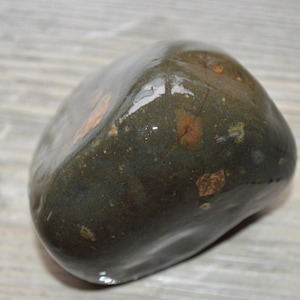 May include: A smooth, polished, rounded stone with a dark olive green base color. The surface features small, irregular patches of reddish-brown and tan mineral inclusions scattered across its glossy, reflective, and slightly uneven texture.