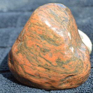Op de afbeelding: Een gepolijste, afgeronde steen met een levendige mix van gebrand oranje en diep houtskoolzwarte wervelende patronen. Het oppervlak is glad en reflecterend en toont natuurlijke minerale variaties met een glanzende afwerking over de onregelmatige vorm.