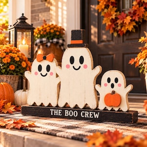 May include: A wooden decoration featuring three white ghost figures standing on a black base with the text THE BOO CREW. The left ghost wears an orange bow, the center ghost wears a black top hat, and the right ghost holds an orange pumpkin.