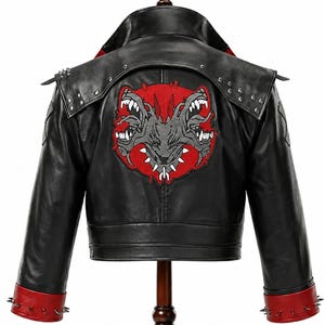 Op de afbeelding: Een zwarte jas met een grote rood-grijze geborduurde patch van een driekoppig wezen op de rug. De jas is voorzien van zilverkleurige metalen studs op de schouderflappen en manchetten, met rode accenten op de manchetten en de kraag.