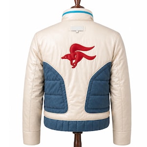 Könnte beinhalten: Eine cremefarbene Jacke mit einem roten, gestickten Fuchsemblem auf der Rückseite. Die Jacke verfügt über blaue, gesteppte Einsätze am unteren Rücken und an den Seiten, einen Stehkragen mit blauem Rand und einen rechteckigen Aufnäher unter dem Kragen.
