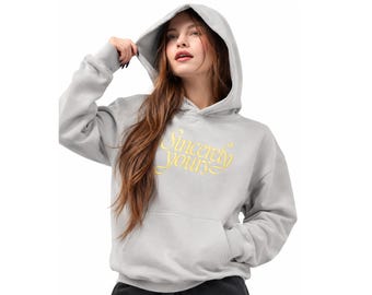 May include: A light gray hooded sweatshirt featuring a front kangaroo pocket and the text "Sincerely yours" printed in a yellow cursive script across the chest. The garment has long sleeves with ribbed cuffs and a matching gray hood.