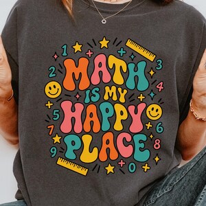 May include: A charcoal grey short-sleeve t-shirt featuring the text "MATH IS MY HAPPY PLACE" in colourful, wavy bubble letters. The design includes small graphics of rulers, smiley faces, stars, and numbers scattered around the central text.