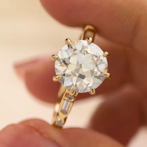 Puede incluir: Un anillo de oro con una gran piedra central redonda de talla brillante sujeta por seis garras. La banda está adornada con piedras más pequeñas de talla baguette rectangular a los lados. El logotipo Everstone JewelryCo es visible en la esquina.