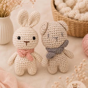 May include: Two small crocheted stuffed animals, a cream-colored rabbit with long ears and a light brown bear with rounded ears. The rabbit wears a small pink scarf, and the bear wears a grey scarf. Both have black bead eyes and stitched noses.