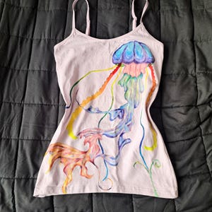 Könnte beinhalten: Ein hellrosa Trägertop mit einer handgemalten Qualle. Die Qualle hat einen blau-violetten Schirm mit fließenden, mehrfarbigen Tentakeln in Orange, Gelb, Rosa und Blau, die sich bis zum unteren Saum des Kleidungsstücks erstrecken.