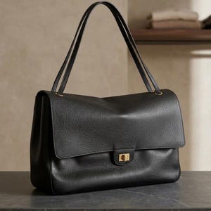 Puede incluir: Un bolso de hombro de cuero negro con textura granulada, que presenta una solapa rectangular grande, dos correas finas para el hombro y un cierre giratorio rectangular de tono dorado centrado en la parte frontal.