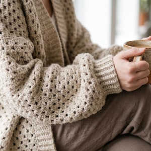May include: A beige crochet cardigan featuring a granny square pattern with long sleeves and ribbed cuffs, paired with brown trousers and a speckled ceramic mug held in both hands, showcasing a textured knit construction.