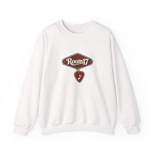 Puede incluir: Una sudadera blanca de manga larga con cuello redondo y un gráfico en el pecho. El diseño muestra un letrero granate en forma de diamante con el texto "Room17" y una púa de guitarra granate colgando, que incluye una nota musical blanca.
