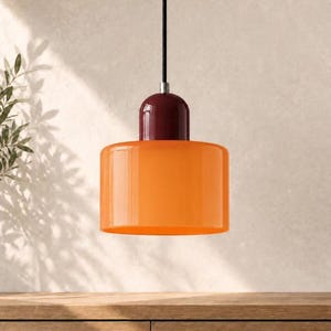 May include: A pendant light fitting featuring a cylindrical orange glass shade suspended by a black cord. The top of the shade is capped with a smooth, rounded, deep burgundy dome component that connects to the black suspension cable.