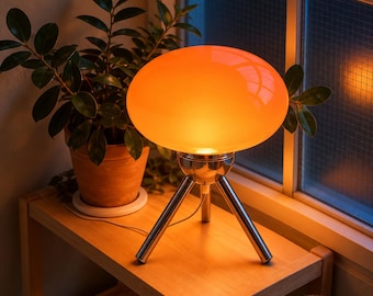May include: A table lamp featuring a smooth, rounded orange glass shade that emits a warm glow. The lamp is supported by three slender, polished silver-toned metal legs arranged in a tripod configuration, providing a stable base for the light fixture.