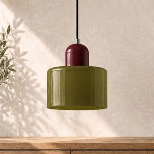 May include: A pendant light fitting featuring a cylindrical olive green glass shade topped with a glossy dark red dome-shaped cap. The lamp is suspended by a thin black cord and includes a small silver-toned metal connector piece.