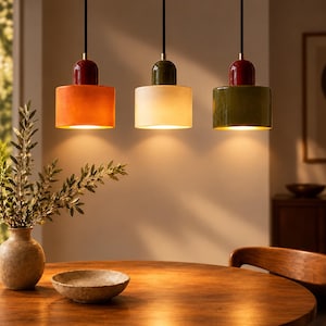 May include: Three cylindrical pendant lights hang from black cords. The left light is orange with a red top, the centre light is cream with a green top, and the right light is green with a red top. Each fixture emits a warm downward glow.
