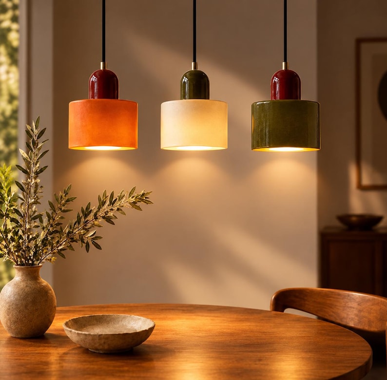 May include: Three cylindrical pendant lights hang from black cords. The left light is orange with a red top, the centre light is cream with a green top, and the right light is green with a red top. Each fixture emits a warm downward glow.