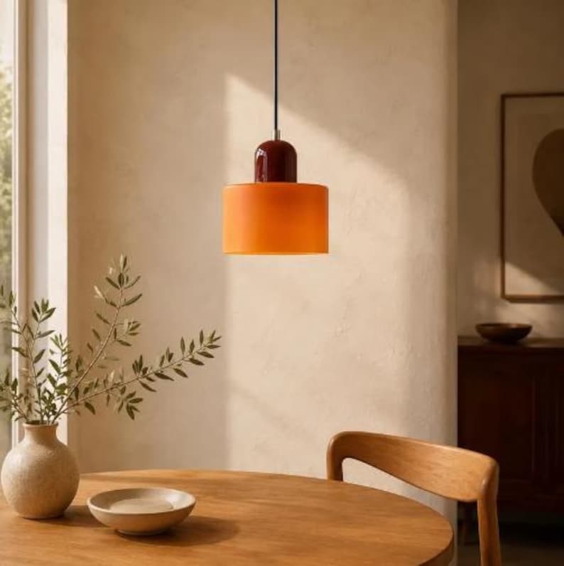 May include: A pendant light fitting featuring a cylindrical orange glass shade suspended by a black cord. The top of the fitting has a smooth, rounded dark red cap that connects to the cord, creating a two-toned colour contrast.