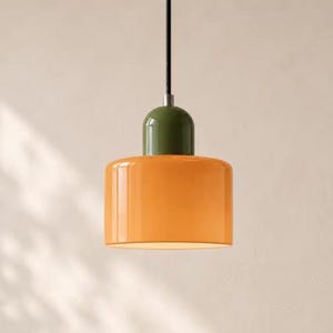 May include: A pendant light fitting featuring a cylindrical orange glass shade topped with a smooth, rounded olive green cap. The lamp is suspended by a thin black cord and includes a silver-toned metal connector at the top of the shade.