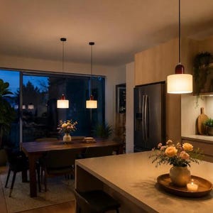 May include: Three cylindrical pendant lights with white frosted glass shades and dark red caps suspended by thin black cords. The lights are positioned over a wooden dining table and a kitchen island, casting a warm glow in the room.