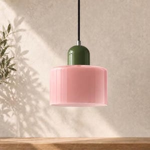 May include: A pendant light fitting featuring a cylindrical, glossy light pink glass shade attached to a smooth, rounded olive green cap. The fitting is suspended by a thin black cord and includes a small silver metal connector piece.