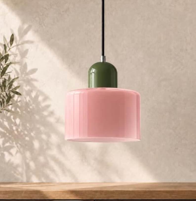 May include: A pendant light fitting featuring a cylindrical, glossy light pink glass shade attached to a smooth, rounded olive green cap. The fitting is suspended by a thin black cord and includes a small silver metal connector piece.