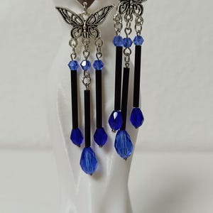 Könnte beinhalten: Ein Paar Ohrhänger mit silberfarbenen Schmetterlingsanhängern an rautenförmigen Steckern. Das Design umfasst hängende silberne Ketten, die mit kleinen blauen Kristallperlen, langen schwarzen zylindrischen Perlen und größeren blauen tropfenförmigen Kristallen verziert sind.