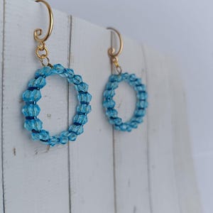 Könnte beinhalten: Ein Paar Ohrringe mit hellblauen, facettierten Kristallperlen in kreisförmiger Anordnung. Die Ohrringe hängen an goldfarbenen Metallhaken, die an der Oberseite ein geschwungenes Spiraldesign aufweisen und mit den Perlenreifen verbunden sind.