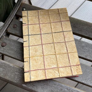 May include: A rectangular notebook with a textured, light tan, fibrous paper cover. The cover features a grid pattern created by thin, intersecting vertical and horizontal threads in black, red, and yellow. The left edge is bound with visible stitching.