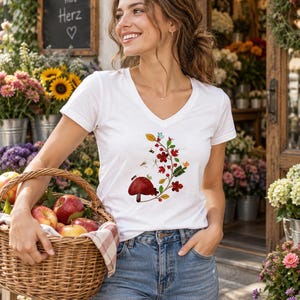 Könnte beinhalten: Ein weißes T-Shirt mit V-Ausschnitt und einem grafischen Aufdruck eines roten Pilzes, kleiner roter Blüten, grüner Blätter und winziger Insekten. Das Oberteil wird mit einer blauen Jeans kombiniert und ein geflochtener Korb mit roten Äpfeln gehalten.