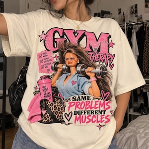 Könnte beinhalten: Ein cremefarbenes T-Shirt mit einer Grafik einer Puppe, die Hanteln hebt. Der Text lautet GYM THERAPY, HOT GIRLS LIFT HEAVY THINGS und SAME PROBLEMS DIFFERENT MUSCLES. Eine Checkliste enthält BAD MOOD, HEAVY WEIGHTS und BETTER ME, neben einer Leoparden-Tasche.