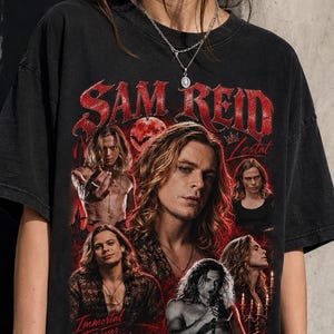 May include: A black short-sleeve t-shirt featuring a red graphic print of Sam Reid. The design includes multiple portraits of the individual, the text "SAM REID", "Lestat", and "Immortal Forever Beautiful", along with a moon and cross symbol.